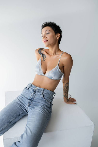 young tattooed woman in blue jeans and satin bra posing on cube on grey background - Foto, imagen