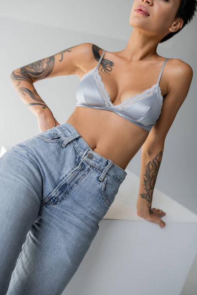 partial view of sexy woman in blue silk bralette and jeans posing with hand on hip on grey background - Valokuva, kuva