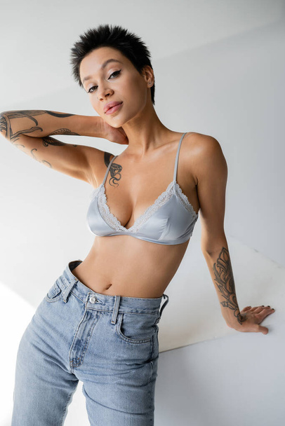 seductive tattooed woman in blue bra and jeans posing near cube with hand behind neck on grey background - Fotoğraf, Görsel