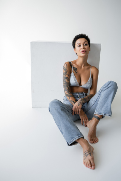 barefoot tattooed woman in blue jeans and bra sitting near cube and looking at camera on grey background - Photo, Image