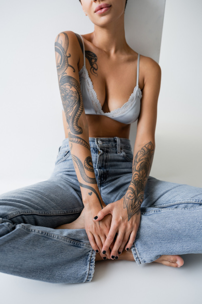 μερική άποψη του τατουάζ ξυπόλητη γυναίκα σε τζιν και σουτιέν κάθεται με σταυρωμένα πόδια σε γκρι φόντο - Φωτογραφία, εικόνα