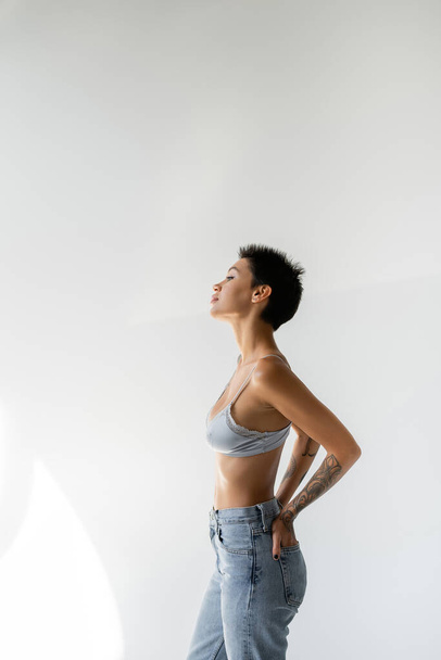 side view of brunette short haired woman in blue bra standing with hands in back pockets of jeans on grey background - Foto, imagen