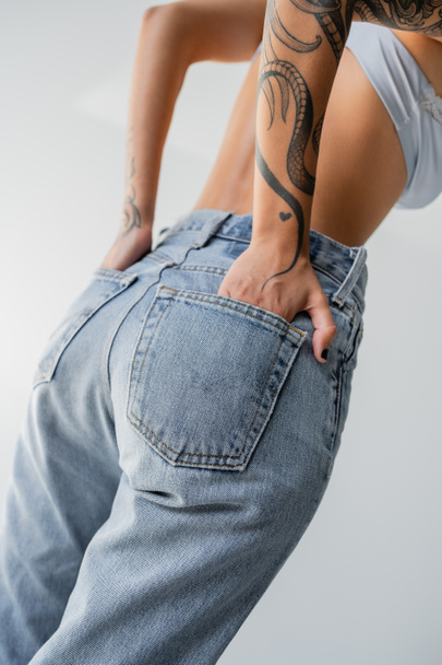 cropped view of tattooed woman in bra posing with hands in back pockets of blue jeans isolated on grey - Foto, Imagem