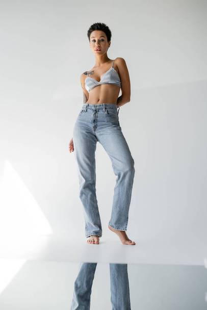 full length of barefoot brunette woman in jeans and bralette standing near mirror on floor on grey background - Photo, image