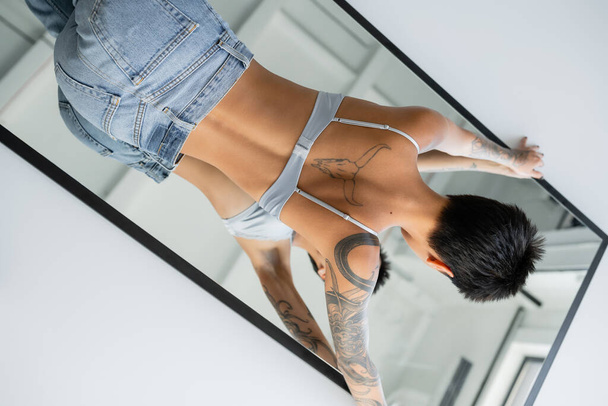 mujer de pelo corto tatuada en sujetador y jeans acostados en el espejo sobre fondo gris - Foto, imagen