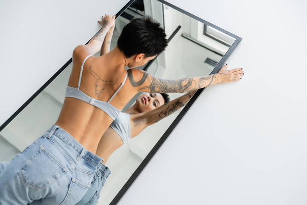 high angle view of seductive tattooed woman in bra and jeans lying on mirror on grey background - Photo, Image