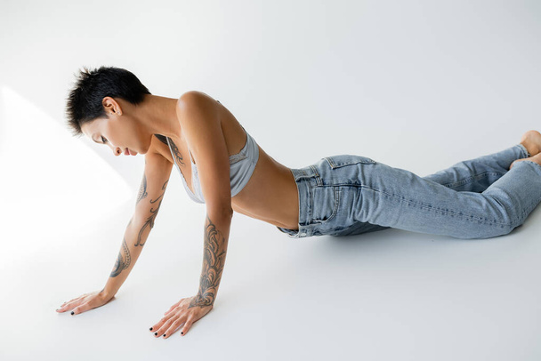 sensual tattooed woman in satin bra and blue jeans posing on grey background - Foto, Bild