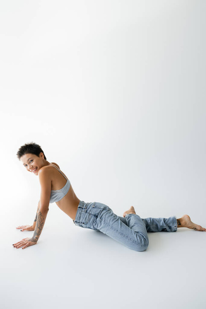 full length of cheerful barefoot woman in bra and jeans looking at camera while posing on grey background - Photo, image