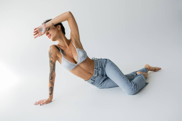 full length of sexy barefoot woman in silk bralette and blue jeans obscuring face with hand while posing on grey background - Fotó, kép