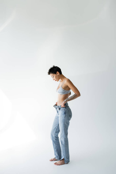 полная длина босиком и стройная женщина в шелковом бюстгальтере и расстегнутых джинсах стоя на сером фоне - Фото, изображение