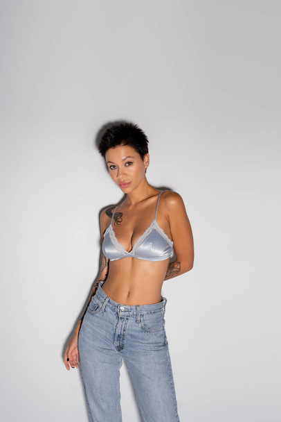 tattooed brunette woman in silk bralette and blue jeans standing with hand behind back on grey background - Foto, afbeelding