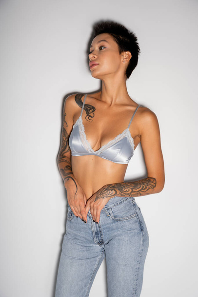 сексуальна татуйована жінка в шовковому бюстгальтері і джинсах дивиться в сторону сірого фону
 - Фото, зображення