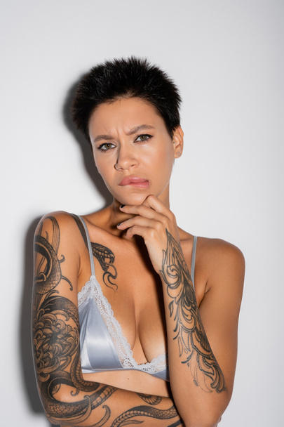 displeased tattooed woman in blue satin bra biting lip and touching chin while looking at camera on grey background - Foto, Imagen