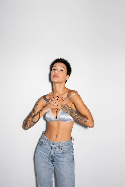 sexy tattooed woman in silk bra and jeans holding hands on chest while looking at camera on grey background - 写真・画像