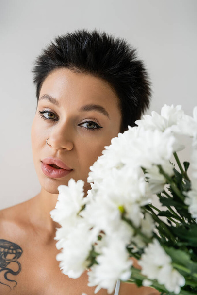 portrait of young woman with piercing and makeup looking at camera near white chrysanthemums isolated on grey - Foto, immagini