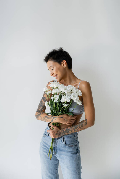 szczęśliwy tatuaż kobieta w biustonosz i dżinsy gospodarstwa bukiet białe kwiaty izolowane na szary - Zdjęcie, obraz