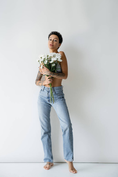 full length of barefoot woman in blue jeans holding bouquet of white chrysanthemums on grey background - Photo, Image