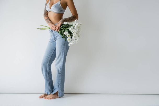 cropped view of barefoot woman in jeans and bra holding white flowers on grey background - Foto, afbeelding