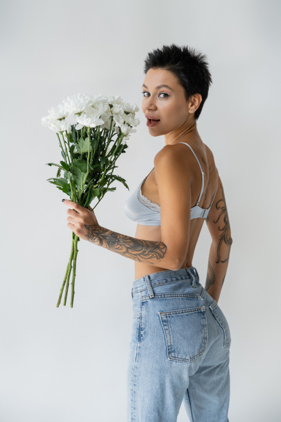 amazed woman in bralette and blue jeans holding bouquet of white flowers and looking at camera isolated on grey - Foto, afbeelding