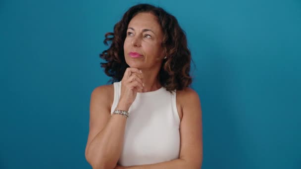 Frau mittleren Alters steht mit zweifelndem Gesichtsausdruck vor isoliertem blauen Hintergrund - Filmmaterial, Video