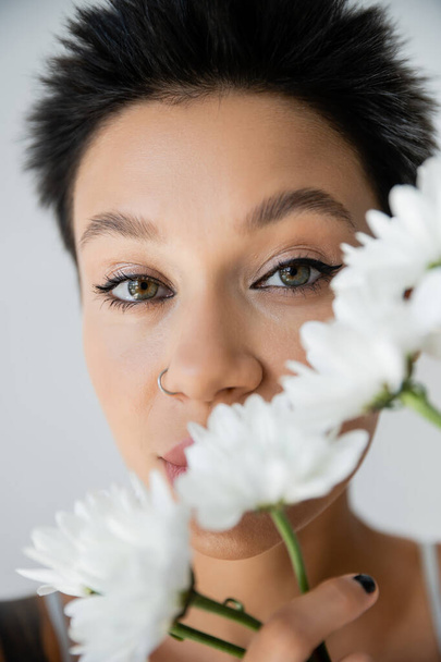 close up portrait of brunette woman with makeup and piercing looking at camera near white flowers isolated on grey - Foto, immagini