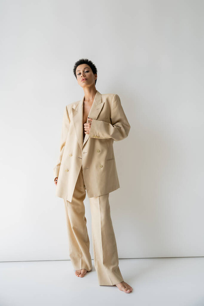 full length of barefoot woman in elegant oversize suit standing and looking at camera on grey background - Foto, afbeelding