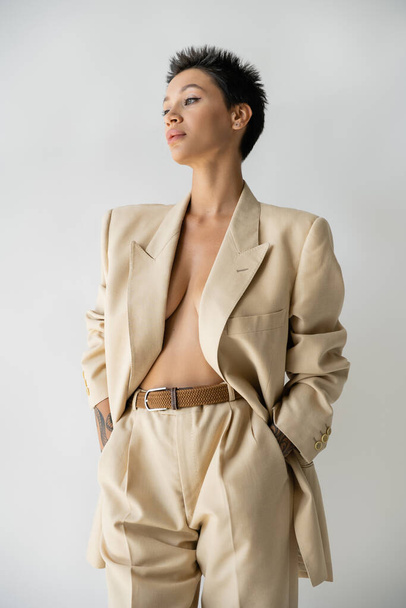 seductive woman in oversize blazer on shirtless body standing with hands in pockets and looking away isolated on grey - Foto, Imagem