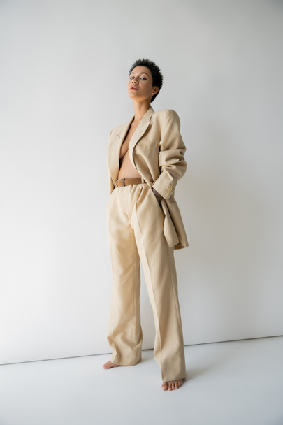 full length of barefoot and shirtless woman in oversize pantsuit standing with hand in pocket on grey background - Photo, Image