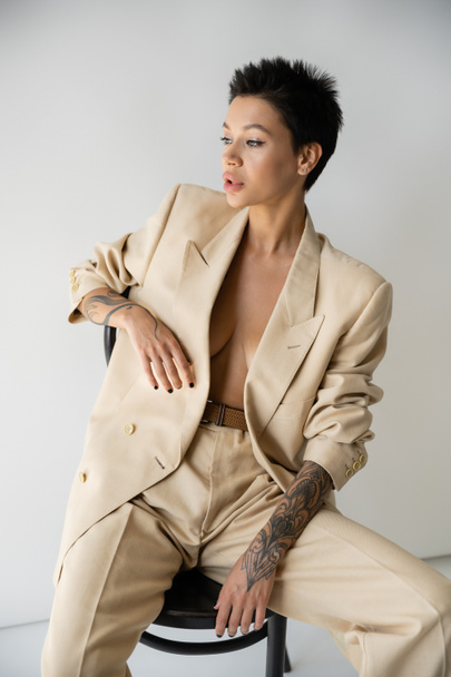 sensual tattooed woman in stylish blazer on shirtless body sitting on chair and looking away on grey background - Photo, Image