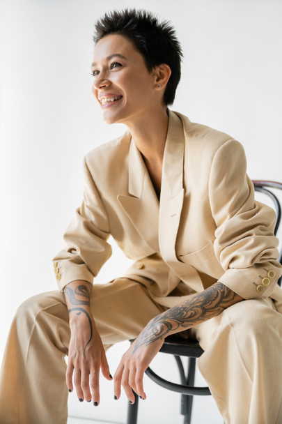 mujer tatuada feliz en traje pantalón de moda sentado en la silla y mirando hacia otro lado sobre fondo gris - Foto, Imagen