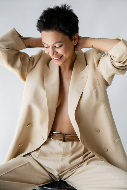 smiling woman in oversize blazer on shirtless body posing with hands behind neck isolated on grey - Фото, изображение