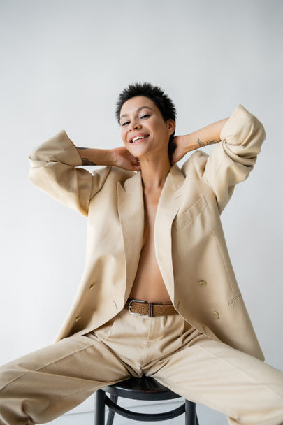 happy shirtless woman wearing blazer on shirtless body and sitting on chair with hands behind neck on grey background - Photo, Image