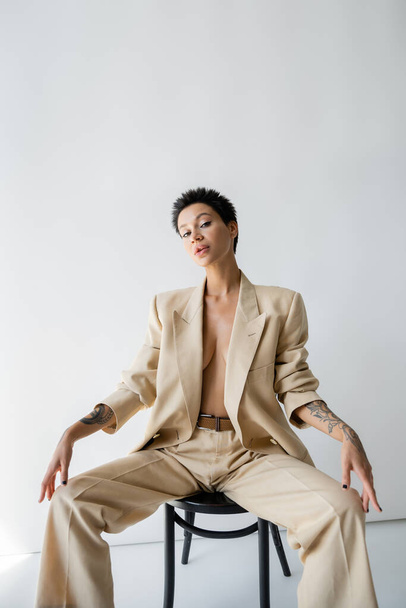 sexy shirtless woman in trendy oversize suit sitting on chair and looking at camera on grey background - Foto, Imagem