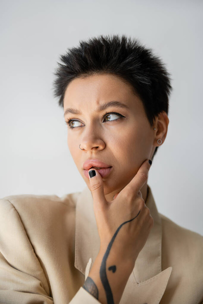 portrait de femme réfléchie en blazer beige touchant lèvre et regardant loin isolé sur gris - Photo, image