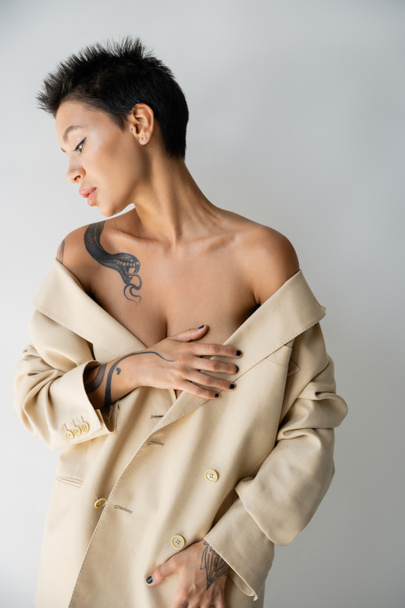 sensual tattooed woman with bare shoulders covering breast with hand while posing in oversize blazer isolated on grey - Фото, зображення