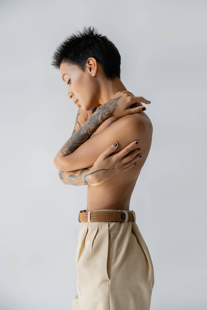 side view of shirtless tattooed woman in trousers embracing herself isolated on grey - Foto, imagen