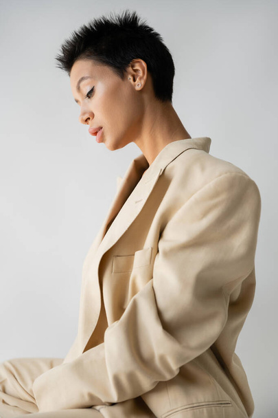side view of stylish brunette woman in oversize blazer sitting isolated on grey - Foto, imagen
