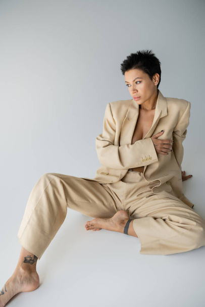 barefoot woman in stylish suit on shirtless body covering breast with hand while sitting on grey background - Foto, Imagen