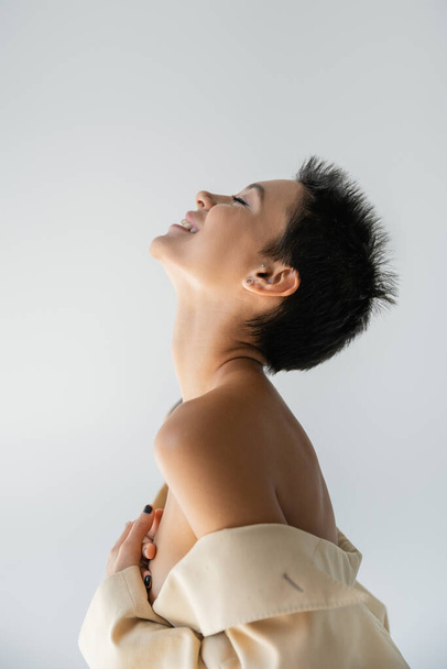 side view of happy and sexy woman in beige blazer with bare shoulders covering breast with hands isolated on grey - 写真・画像