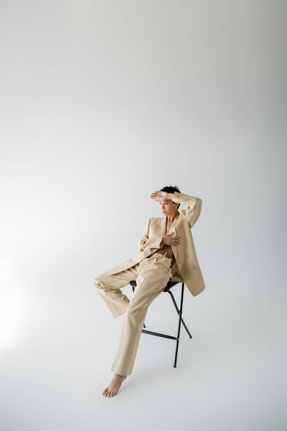 full length of barefoot woman in beige suit sitting on chair and holding hand near forehead on grey background - Fotografie, Obrázek
