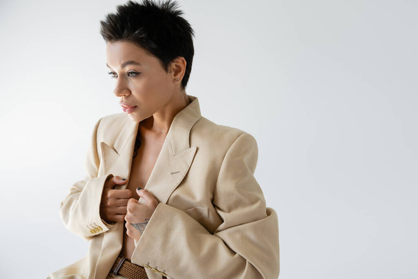 young woman with short brunette hair wearing beige blazer on shirtless body and looking away isolated on grey - Photo, Image
