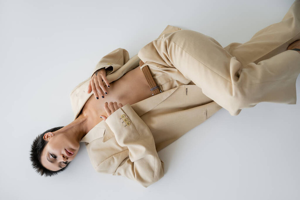 верхний вид женщины без рубашки в модном брючном костюме, прикрывающей грудь руками, лежащей на сером фоне - Фото, изображение