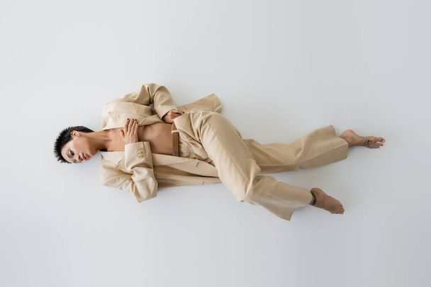 top view of barefoot and shirtless woman in beige suit covering breast with hand while lying on grey background - Fotó, kép