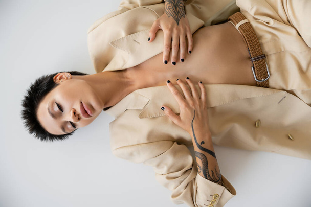 top view of sexy woman in beige blazer covering breast with hands while lying with closed eyes on grey background - Foto, Imagen