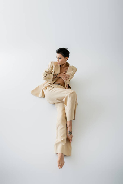 magas szög kilátás barna nő divatos öltöny borító mell kézzel, miközben ül, és nézi el szürke háttér - Fotó, kép