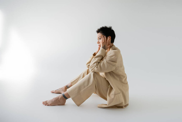 side view of tattooed barefoot woman in beige pantsuit sitting with hands near face on grey background - Foto, Imagem