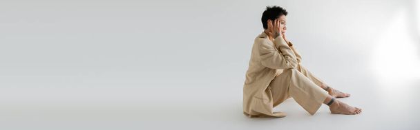 vista laterale della giovane donna bruna in abito alla moda seduta con le mani vicino alla testa su sfondo grigio, banner - Foto, immagini