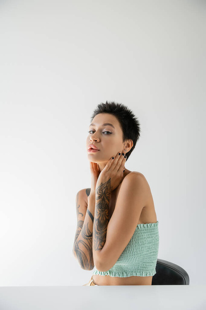 young tattooed woman in strapless top holding hands near neck while sitting at table on grey background - Fotó, kép