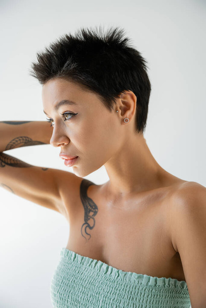 young tattooed woman in strapless top holding hand near head and looking away isolated on grey - Foto, immagini