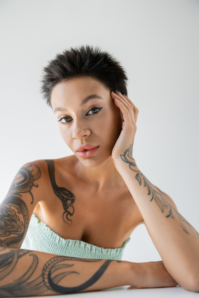πορτρέτο της γυναίκας με τατουάζ σε στράπλες πάνω μέρος ποζάροντας με το χέρι κοντά στο πρόσωπο απομονωμένο σε γκρι - Φωτογραφία, εικόνα